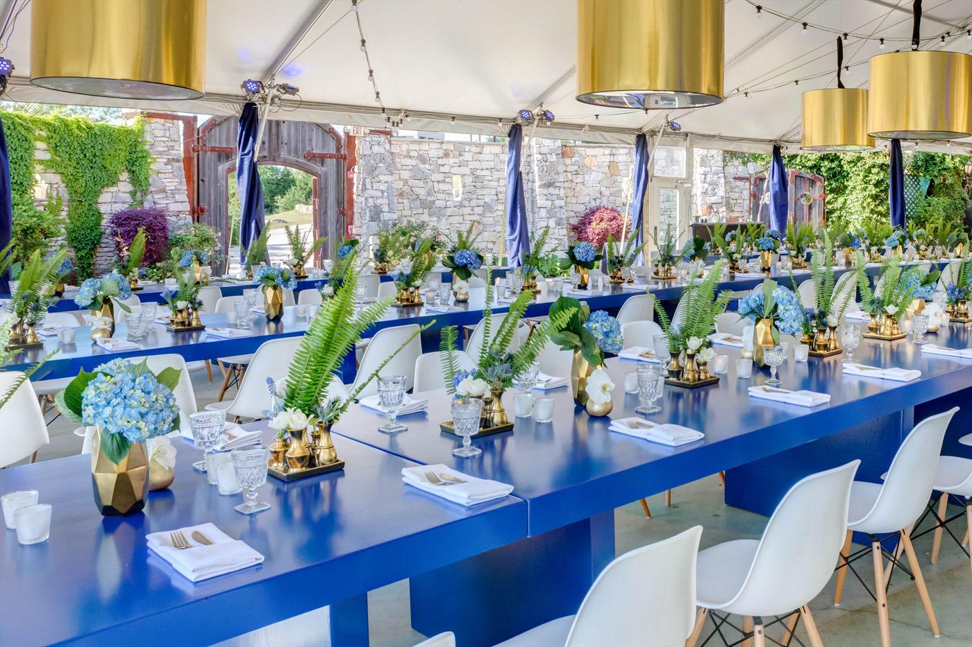 Long cobalt-blue banquet tables under a white tent, set with white linens, gold vessels, blue hydrangeas and ferns, with brass pendant lights overhead.